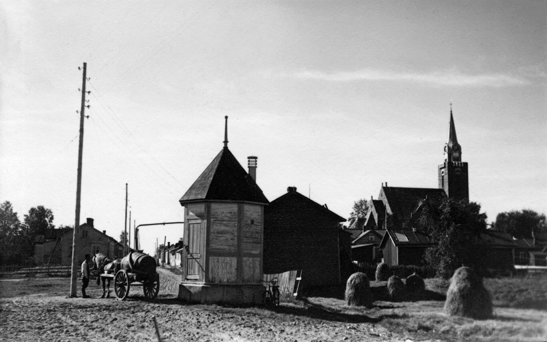 Vanha mustavalkoinen valokuva Jaakopinlähteestä Reiponkadulla Wanhassa Raahessa. Kuvassa näkyy hevonen kärryineen, heinäpaaleja sekä taustalla Raahen kirkko.